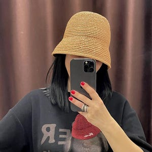 May include: A person wearing a black t-shirt with a graphic of a teddy bear wearing a red hat. The text "Good" is printed on the shirt. The person is holding a black smartphone and wearing a straw bucket hat.