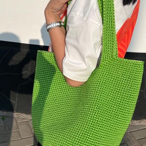 May include: A vibrant green, hand-crocheted tote bag with a wide strap. The bag is made of textured yarn and has a simple, open design. The bag is held by a person wearing a white shirt.