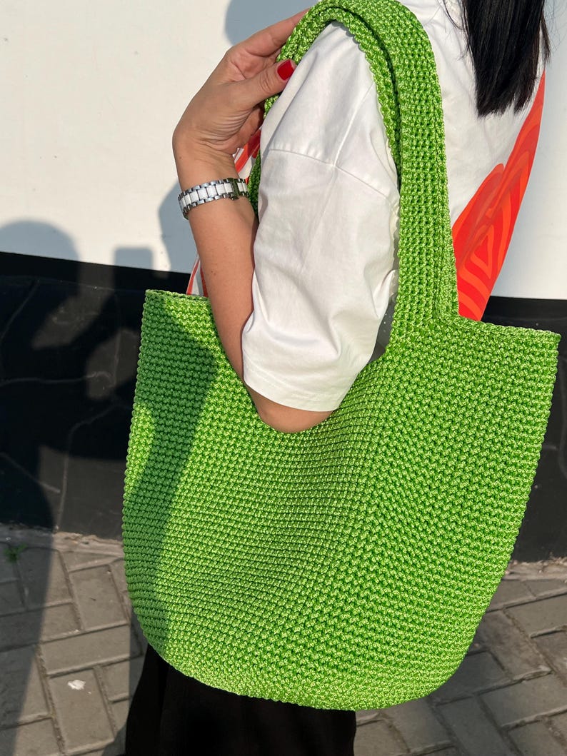 May include: A vibrant green, hand-crocheted tote bag with a wide strap. The bag is made of textured yarn and has a simple, open design. The bag is held by a person wearing a white shirt.