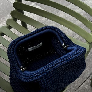 May include: Open navy blue crochet handbag with a structured shape. The bag features a textured stitch pattern and a dark blue interior. The metal clasp is open, revealing the inside. The bag is sitting on a green chair.