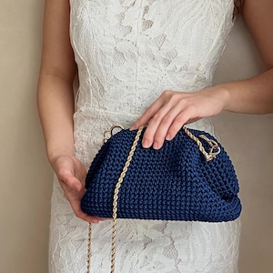 May include: A navy blue crocheted handbag with a gold chain strap. The bag is held by a person wearing a white lace dress.