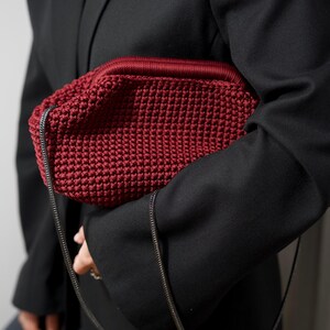 May include: A red crocheted bag with a black chain strap. The bag is being held by a person wearing a black blazer.