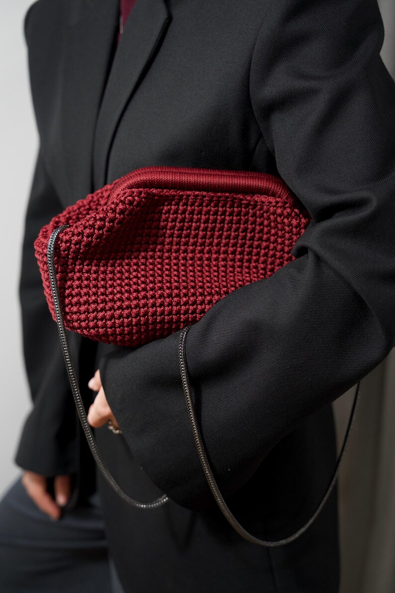 May include: A red crocheted bag with a black chain strap. The bag is being held by a person wearing a black blazer.
