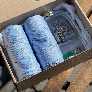 Può includere: Due bobine di cordoncino blu chiaro, un sacchetto di plastica trasparente con ferramenta metallica e anelli di plastica verde sono all'interno di una scatola marrone.