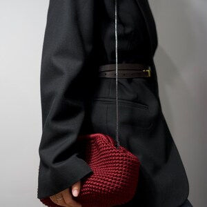 May include: A woman wearing a black blazer with a brown belt and a dark red crocheted bag.