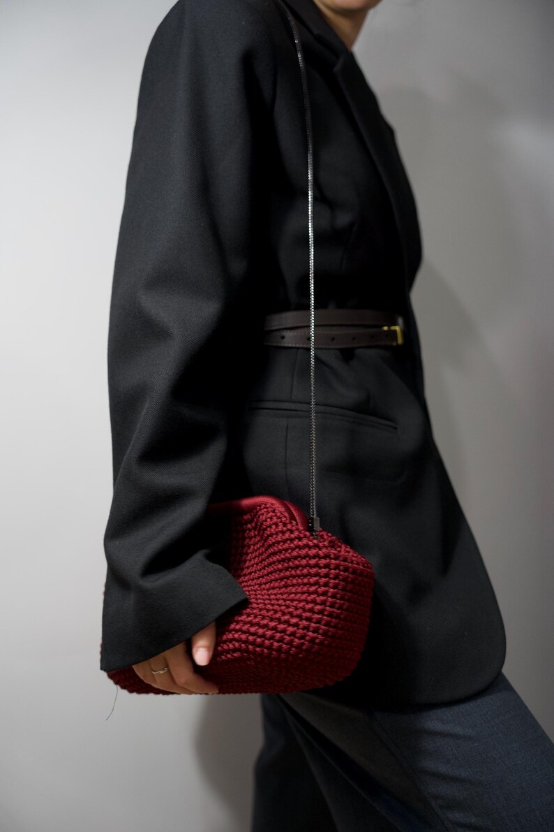 May include: A woman wearing a black blazer with a brown belt and a dark red crocheted bag.