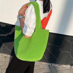 May include: A vibrant green, hand-crocheted tote bag with a rectangular form and strong handles. The bag is made from thick yarn, and the stitch pattern is visible. The bag is carried over the shoulder.