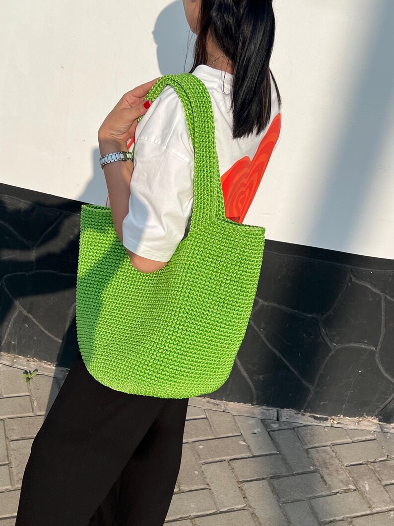 May include: A vibrant green, hand-crocheted tote bag with a rectangular form and strong handles. The bag is made from thick yarn, and the stitch pattern is visible. The bag is carried over the shoulder.