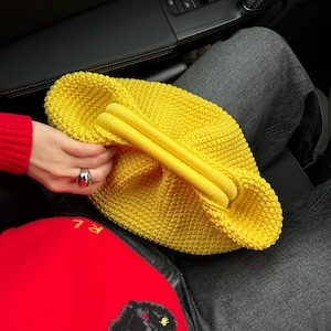 May include: A bright yellow crocheted bag with a unique, gathered design. The bag is made of thick yarn and has a soft, textured feel.