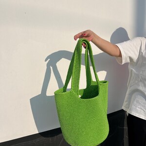 May include: A vibrant green crochet tote bag with long handles. The bag is held up against a white wall with a shadow cast on the wall. The bag is a solid, bright green colour.