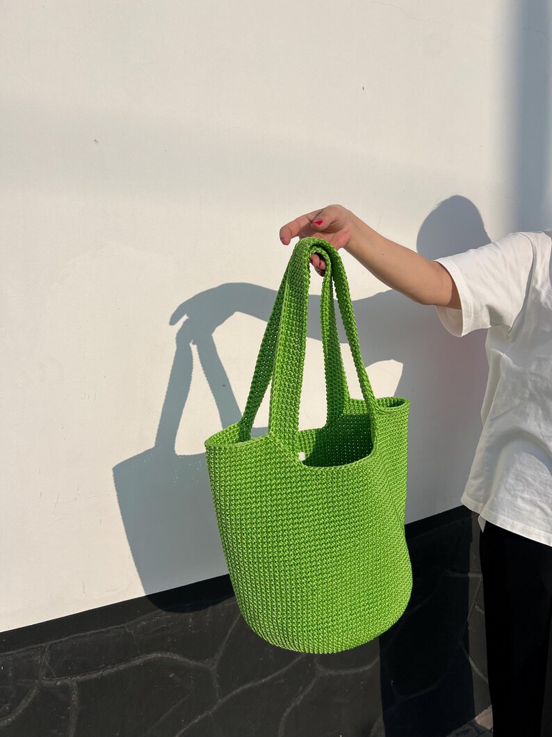 May include: A vibrant green crochet tote bag with long handles. The bag is held up against a white wall with a shadow cast on the wall. The bag is a solid, bright green colour.