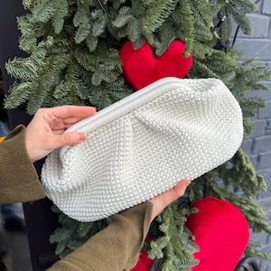 May include: A light gray, crocheted clutch with a zipper closure. The bag is held in front of a backdrop of evergreen branches and a red heart-shaped decoration. The clutch has a rounded shape and a textured surface.
