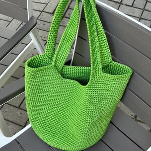 May include: A vibrant green, crocheted tote bag with two long handles. The bag is made of a textured yarn and has a rounded base. The bag is resting on a grey and white chair.