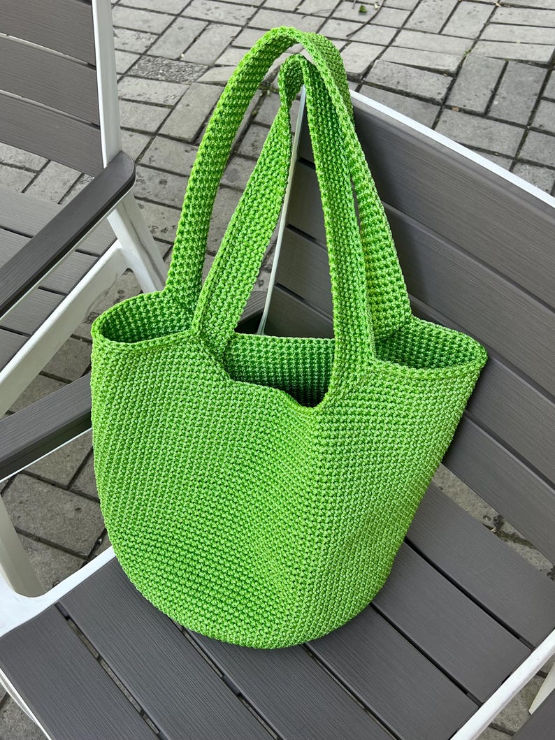 May include: A vibrant green, crocheted tote bag with two long handles. The bag is made of a textured yarn and has a rounded base. The bag is resting on a grey and white chair.