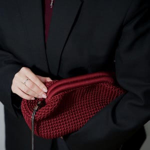 May include: A person wearing a black blazer holds a red crocheted bag with a silver chain strap.