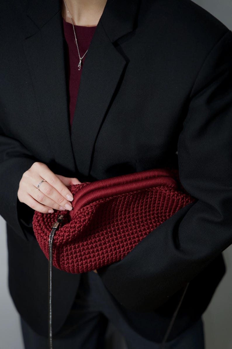 May include: A person wearing a black blazer holds a red crocheted bag with a silver chain strap.