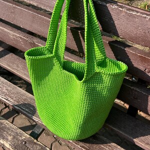 May include: A vibrant green, hand-crocheted tote bag with strong handles. The bag has a rounded shape and a textured appearance, suggesting a durable and stylish accessory. The bag is sitting on a wooden bench.
