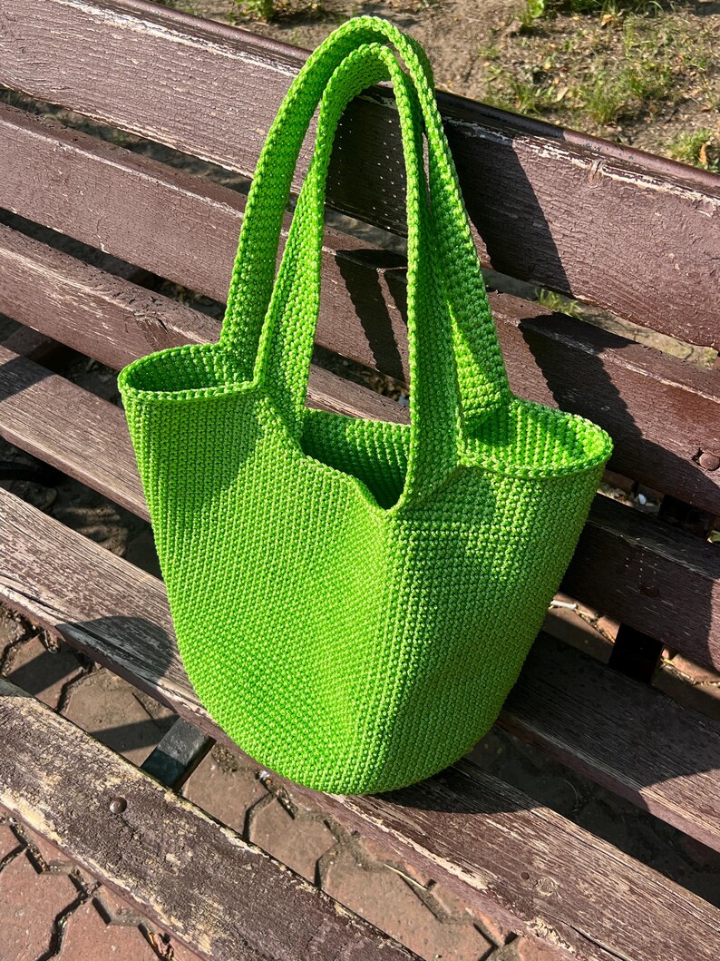 May include: A vibrant green, hand-crocheted tote bag with strong handles. The bag has a rounded shape and a textured appearance, suggesting a durable and stylish accessory. The bag is sitting on a wooden bench.