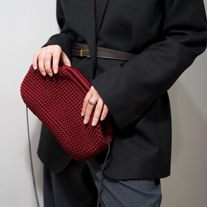 May include: A woman wearing a black blazer with a brown belt and a dark red crocheted bag with a black strap.