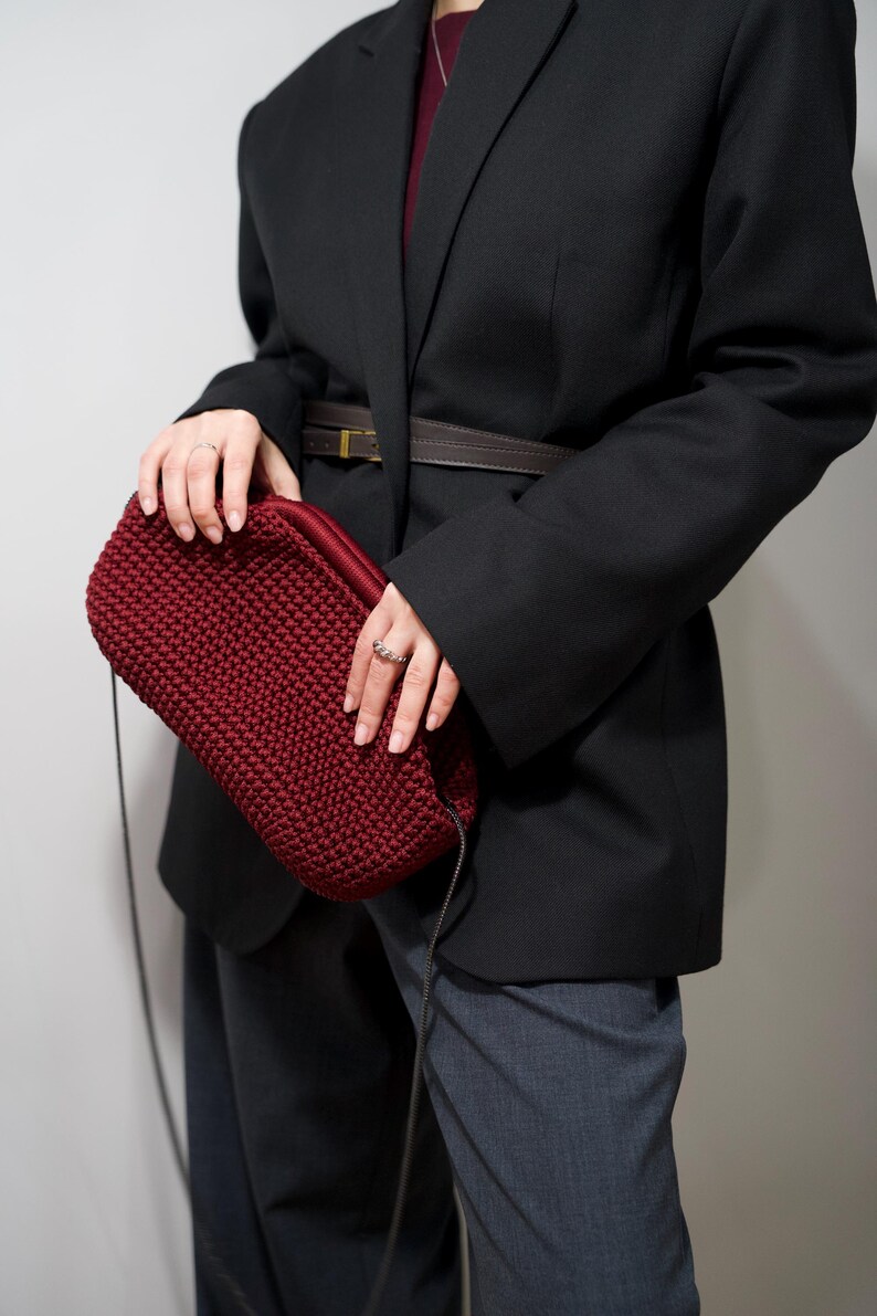 May include: A woman wearing a black blazer with a brown belt and a dark red crocheted bag with a black strap.