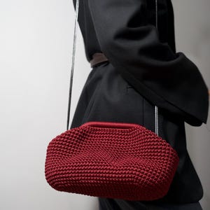 May include: A red crocheted handbag with a silver chain strap. The bag is worn over the shoulder of a person wearing a black blazer.