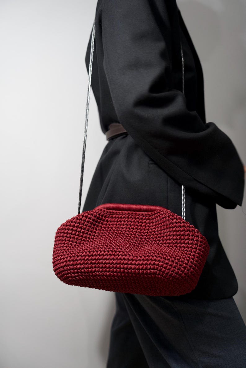 May include: A red crocheted handbag with a silver chain strap. The bag is worn over the shoulder of a person wearing a black blazer.