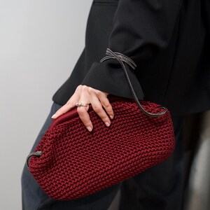May include: A close-up of a red crocheted handbag with a black chain strap. The bag is held by a person wearing a black blazer and dark gray pants.