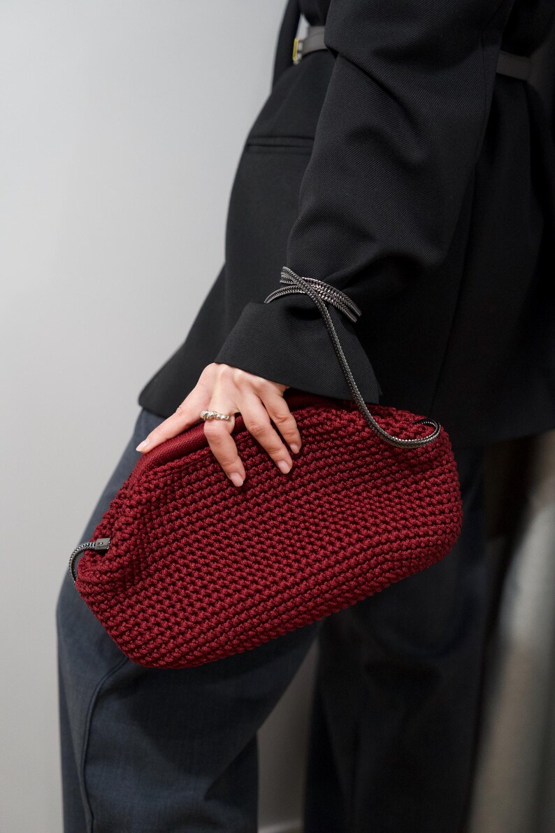 May include: A close-up of a red crocheted handbag with a black chain strap. The bag is held by a person wearing a black blazer and dark gray pants.