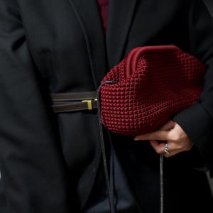 May include: A person wearing a black blazer with a brown leather belt and a dark red knitted bag with a black strap.