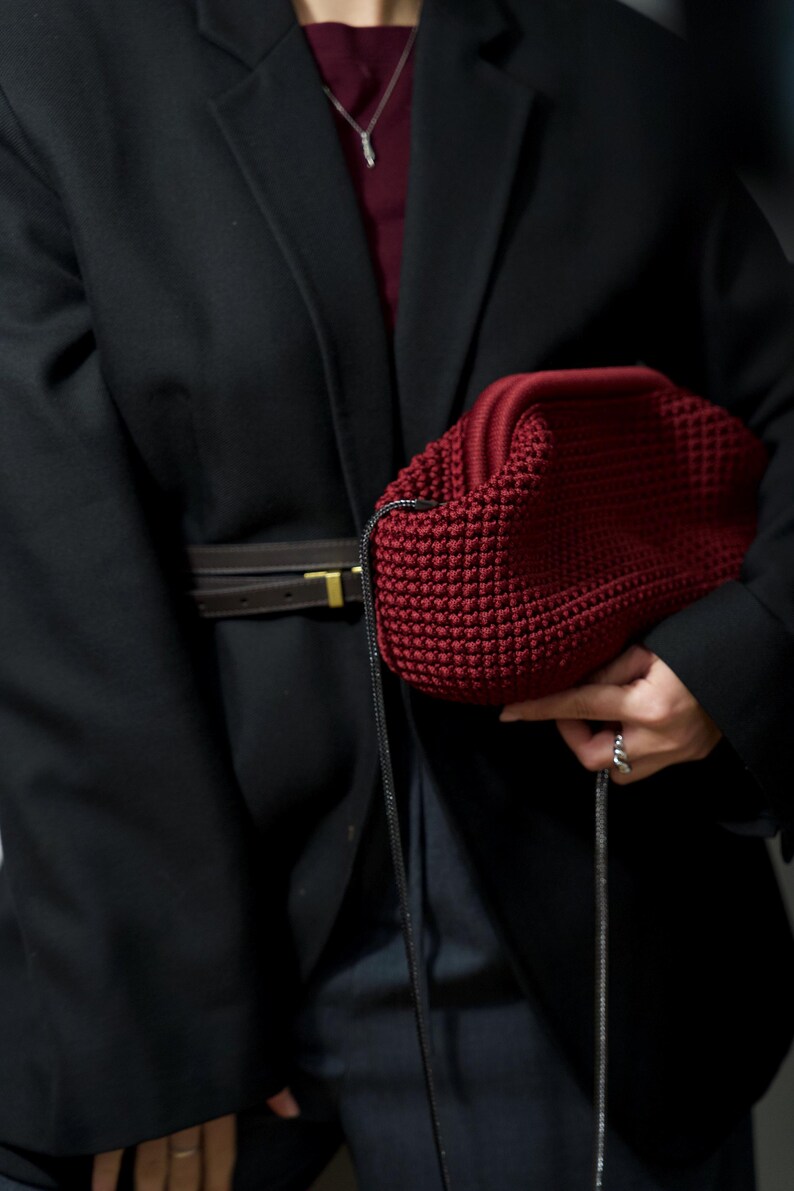May include: A person wearing a black blazer with a brown leather belt and a dark red knitted bag with a black strap.