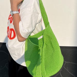 May include: A vibrant green, knitted shoulder bag with a broad strap. The bag has a rounded form and a textured look. The strap is knitted and matches the bag's colour. The bag is held by a person wearing a white t-shirt.