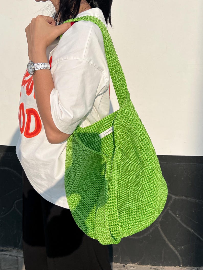 May include: A vibrant green, knitted shoulder bag with a broad strap. The bag has a rounded form and a textured look. The strap is knitted and matches the bag's colour. The bag is held by a person wearing a white t-shirt.