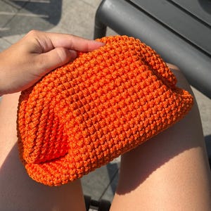 May include: A bright orange clutch bag featuring a dense, textured crochet knit pattern. The pouch has a rounded, soft rectangular shape with a gathered top closure, showcasing a consistent, uniform stitch throughout the entire exterior surface.