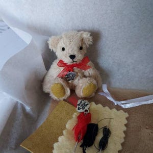 May include: A small, cream-colored teddy bear with a red ribbon and a heart-shaped charm that says "Made With Love." The bear has yellow paw pads and is surrounded by craft supplies, including needles and thread. The bear is sitting in a white box.