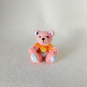 Könnte beinhalten: Ein Miniatur-Teddybär aus weichem, rosafarbenem Plüschstoff. Der Bär hat ein leuchtend orangefarbenes Band um den Hals, kleine schwarze Perlenaugen, eine gestickte schwarze Nase sowie gemusterte Stoffakzente an Ohren und Pfoten.