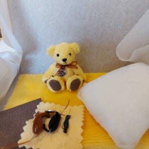 May include: A small yellow teddy bear wearing a brown bow sits on a yellow surface with a brown fabric square and a white pillow. The bear has a small heart-shaped charm with the word "Love" on it.