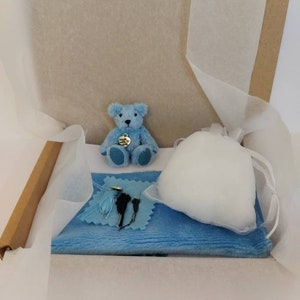 May include: A light blue teddy bear kit inside a cardboard box. The kit includes a small blue teddy bear, a blue felt square with sewing supplies, and a bag of stuffing. The box is lined with white tissue paper.