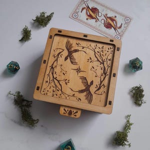 Op de afbeelding: Een houten doos met een scharnierend deksel en een gesneden ontwerp van twee draken die rond een boom vliegen. De doos is gemaakt van licht hout en heeft een natuurlijke afwerking. De draken zijn gesneden in een donkerbruine kleur en de boom is gesneden in een lichtere bruine kleur.