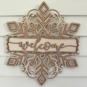 May include: Wooden welcome sign with a layered snowflake design. The sign is painted in a light wood tone and features the word "welcome" in a cursive font.