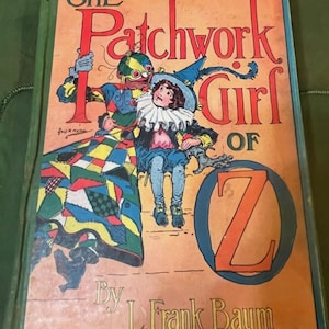 May include: The cover of a vintage children's book titled "The Patchwork Girl of Oz" by L. Frank Baum. The cover features a colorful patchwork girl sitting on a wooden step with a puppet-like figure standing behind her.