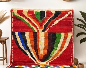 May include: A colorful, abstract rug with a red background and a central design of a stylized tree with green, blue, orange, and yellow stripes. The rug has a fringe border and is laid out on a blue rug.