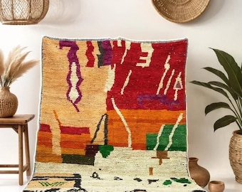 May include: A colorful, abstract patterned rug with a fringe border. The rug is laid out on a dark blue rug, with a wooden side table and a potted plant in the background.