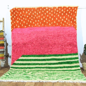 May include: A vibrant, colorful rug with a geometric design. The rug features a top section of orange with white dots, a middle section of bright pink, and a bottom section of green and off-white stripes. The rug has a shaggy texture and a fringed edge.