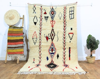 May include: Cream-colored, hand-woven rug with geometric patterns in red, black, and blue. The rug features diamond and triangle shapes, with a fringe along the bottom edge. A straw hat and a colorful striped textile are visible in the background.