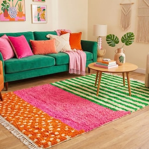 May include: A vibrant living room scene featuring a green velvet sofa with pink and orange pillows. A colorful rug with orange, pink, and green sections sits on the wooden floor. Decorative wall art and a round wooden coffee table complete the space.