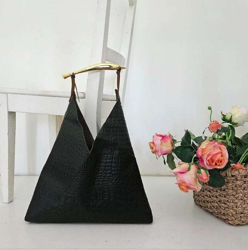 May include: A black leather tote bag with a gold-colored handle. The bag has a triangular shape and a textured surface. The handle is attached with brown leather straps. A bouquet of pink and peach roses sits nearby.