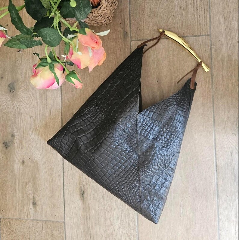 May include: A dark brown, textured leather handbag with a unique triangular shape. The bag features a gold-colored handle and brown leather straps. The bag is placed on a wooden surface, with pink and yellow roses in the background.
