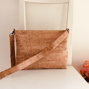 Könnte beinhalten: Rechteckige Umhängetasche aus hellbraunem Kork. Die Tasche hat einen langen, verstellbaren Riemen und einen Klappverschluss. Der Kork hat ein natürliches, strukturiertes Aussehen. Die Tasche steht auf einer weißen Oberfläche.