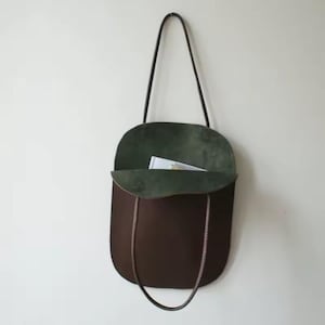 May include: A brown leather tote bag with a dark green interior. The bag has a rounded shape with a long, thin strap and a shorter handle. A book and pen are visible inside the open top.