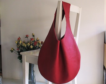 May include: A large, red leather hobo bag hangs on a white chair. The bag has a single shoulder strap and a rounded bottom. The bag is empty. A vase of flowers sits on a small white table to the left of the chair.